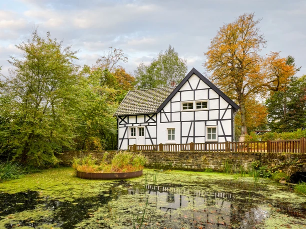 Emscherquellhof, Holzwickede <p>Fachwerkhaus am Teich mit grüner Umgebung, bewölkter Himmel, herbstliche Bäume im Hintergrund.</p>