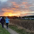 Spiritueller Spaziergang Eine Gruppe von Menschen wandert bei Sonnenuntergang einen Feldweg entlang, umgeben von Feldern.