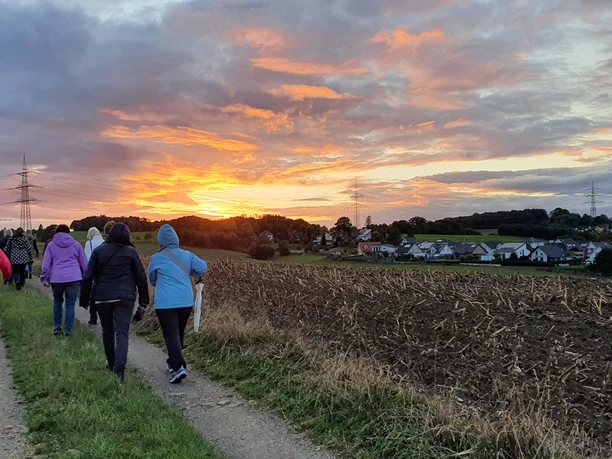 Spiritueller Spaziergang Eine Gruppe von Menschen wandert bei Sonnenuntergang einen Feldweg entlang, umgeben von Feldern.