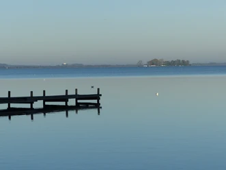 P1040719-2.JPG Ein Steg ragt in einen ruhigen See, während am Horizont eine kleine Insel unter klarem Himmel liegt.