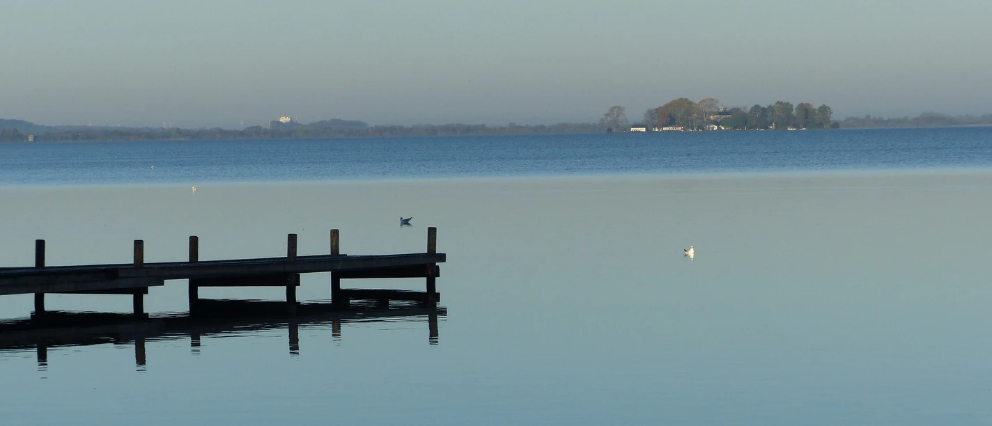 P1040719-2.JPG Ein Steg ragt in einen ruhigen See, während am Horizont eine kleine Insel unter klarem Himmel liegt.