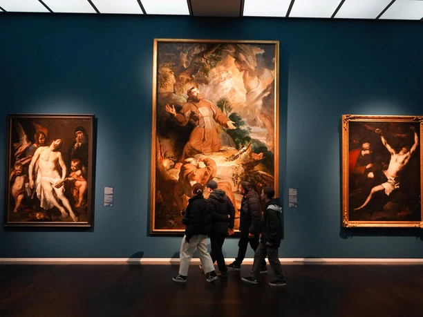 Visitors observe large classical paintings in a dimly lit museum gallery with blue walls.
