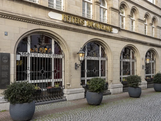 Peters Brauhaus Historische Front von Peters Brauhaus in Köln, geschmückt mit runden Fenstern, Sandsteinfassade sowie Eisenverzierungen. Pflanzkübel säumen den gepflasterten Gehweg.Historic façade of Peters Brauhaus in Cologne, adorned with round windows, sandstone façade and iron decorations. Planters line the paved walkway.