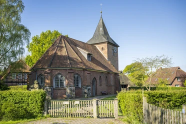 Historic red-brick church with slate roof and clock tower, surrounded by greenery on a sunny day.