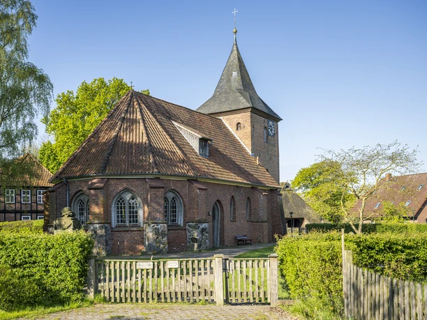 Historic red-brick church with slate roof and clock tower, surrounded by greenery on a sunny day.