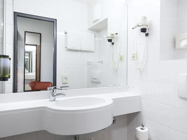 Badezimmer im ACHAT Hotel Monheim am Rhein Helles, modernes Badezimmer mit großem Spiegel, Waschbecken, Haartrockner und Handtuchhalter.
