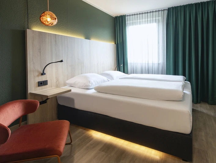Business Doppelzimmer im ACHAT Hotel Monheim am Rhein Doppelzimmer mit modernem Design, geräumigem Bett, schickem Sessel, Holzdetails und eleganter Beleuchtung.