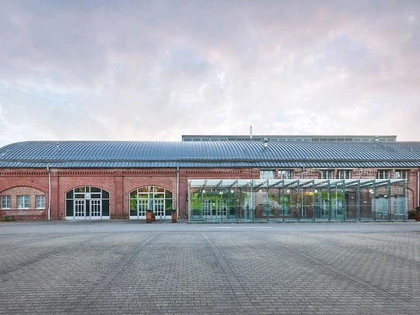 Rohrmeisterei, Schwerte Altes Backsteingebäude mit Glas-Anbau und großem gepflasterten Vorplatz bei bedecktem Himmel.