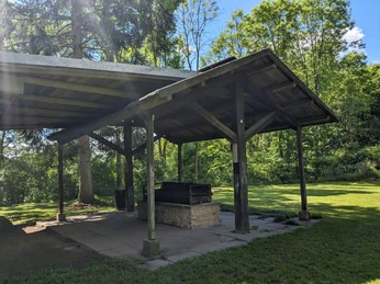 Grillplatz Hube Überdachter Grillplatz mit Steinfeuerstelle auf einer Wiese, umgeben von hohen Bäumen im Sonnenschein.