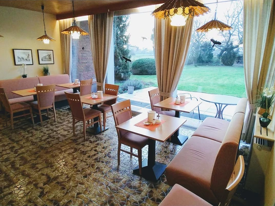 Hotel-Pension Bevertal Garni - Frühstücksraum Frühstücksraum mit Tischgruppen, gepolsterten Stühlen und Sitzbänken, Blick in den grünen Garten, an der Wand ein Holzbuffet und Bilder