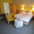 Hotel-Pension Bevertal Garni - Hotelzimmer Doppelzimmer mit Teppichfußboden, Bett und Nachttischen aus Holz, über dem Kopfende Lampen, gelbe Tapete, Polstersessel in senfgelb & kleiner Tisch vor dem Bett platziert, Schiebetürenschrank, Blick in das angrenzende separate Badezimmer