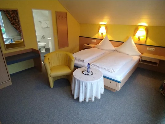 Hotel-Pension Bevertal Garni - Hotelzimmer Doppelzimmer mit Teppichfußboden, Bett und Nachttischen aus Holz, über dem Kopfende Lampen, gelbe Tapete, Polstersessel in senfgelb & kleiner Tisch vor dem Bett platziert, Schiebetürenschrank, Blick in das angrenzende separate Badezimmer