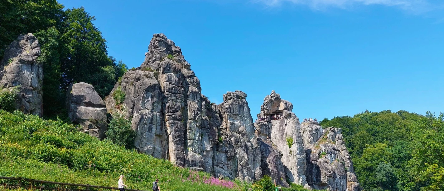 Externsteine_CC_BY_SA_BadSalzuflen_KerstinPaar.jpg Sandsteinfelsen in Norddeutschland, bekannt als Externsteine, umgeben von Wald und Grünflächen.