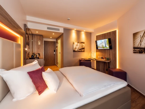 Stadtzimmer im Best Western Plus Hotel Stadtquartier Haan Modernes Hotelzimmer mit Doppelbett, warmem Licht, Flachbildschirm, Holzmöbeln und Schreibtisch.