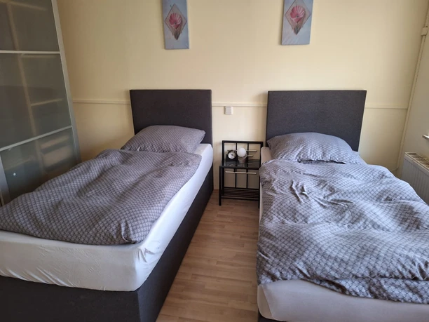 Schlafzimmer in der Ferienwohnung Senjic in Heiligenhaus Zwei nebeneinander stehende Einzelbetten mit grauen Kopfteilen und Bettwäsche, flankiert von einem Nachttisch.