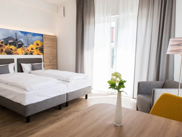 Boardinghouse am Teuto Lengerich_Doppelzimmer (5).jpg Modernes Doppelzimmer mit hellen Wänden, Blumenbild, Schreibtisch, Sessel und großen Fenstern.