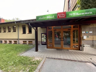 Ingang van een actief hotel met deuren met houten frame en een groen bord met de tekst "Welcome", met een drankautomaat ernaast.