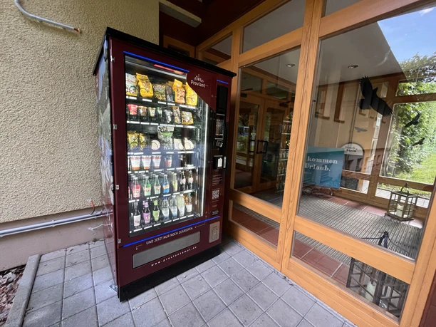 A red vending machine with snacks and drinks stands next to a glass door; in the background you can see a sign saying "Welcome to vacation".