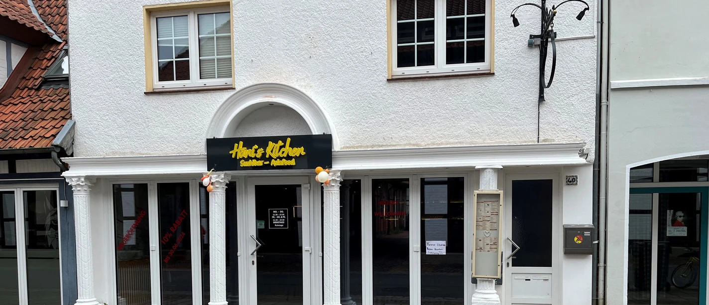 Hani´s Kitchen Das Restaurant Hani’s Kitchen präsentiert eine weiße Fassade mit großen Fenstern und einem gelben Logo.