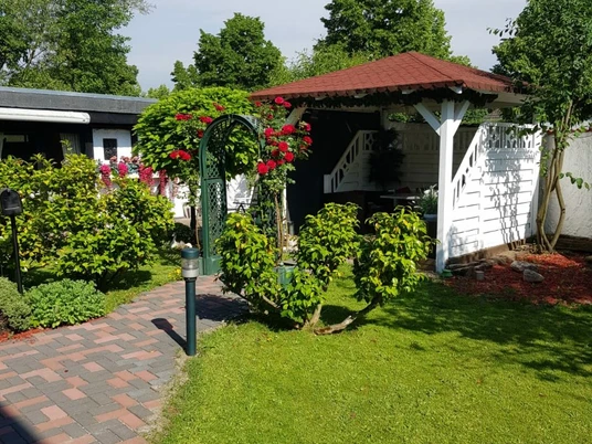 Garten im Hotel am Hallenbad in Ratingen Gepflegter Garten mit gepflastertem Weg, blühenden Rosen an einem Torbogen und einem kleinen Pavillon.