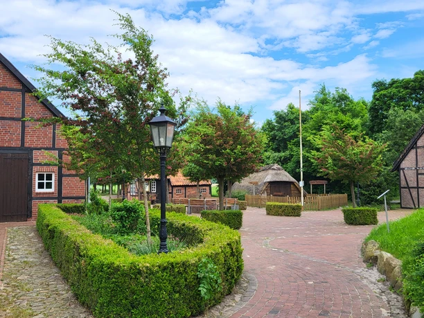 Wippingen, Heimathaus ©Naturpark Hümmling.jpg Historischer Hof mit Fachwerkhäusern, Kopfsteinpflasterweg, grünen Hecken und blühenden Bäumen.