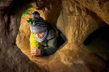 Kind met pet en hoofdlamp kruipt door een smalle, zandkleurige grottunnel. De sfeer is avontuurlijk en gefocust.