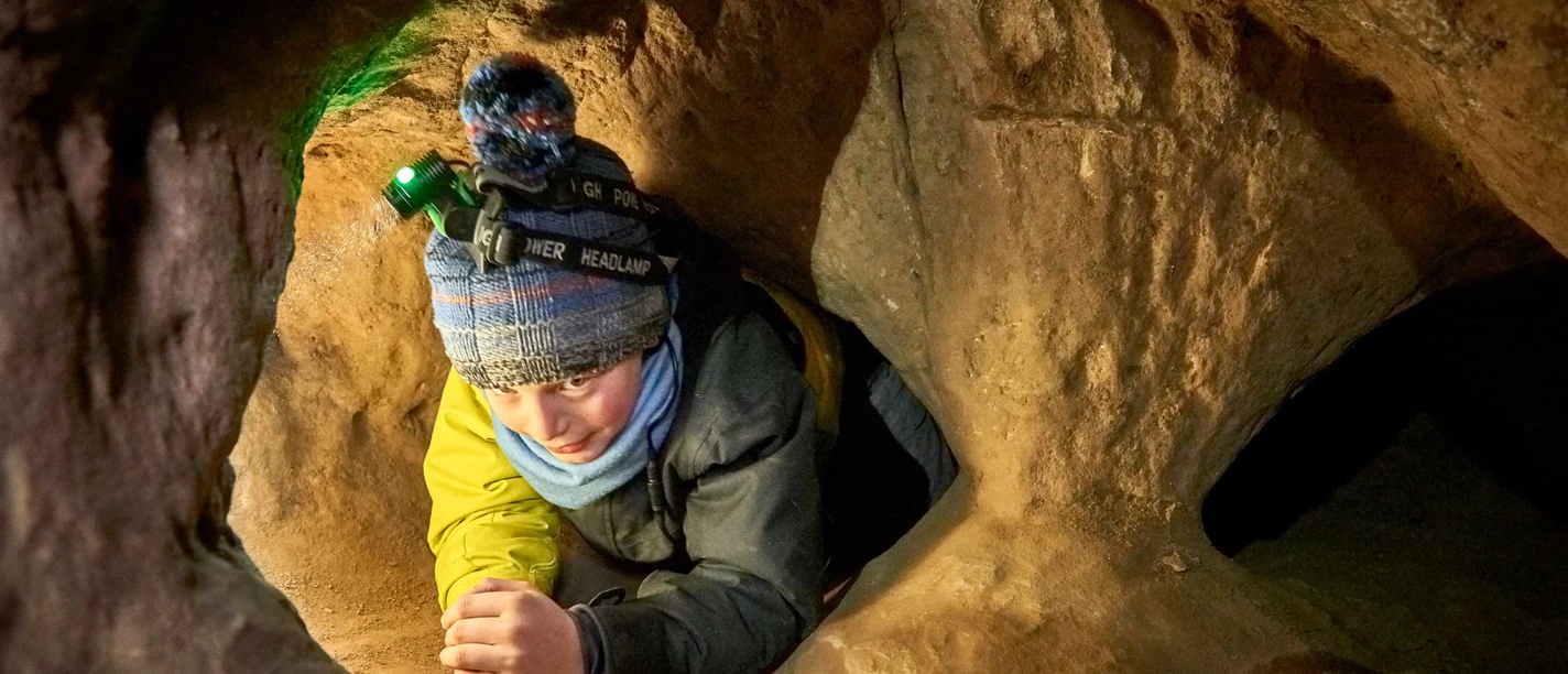 Child with cap and headlamp crawls through a narrow, sand-colored cave tunnel. The atmosphere is adventurous and concentrated.