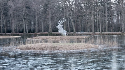 Metallene Hirschskulptur auf kleiner Insel im teils gefrorenen Schlossteich, umgeben von kahlen Bäumen