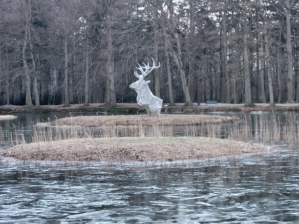 Metallene Hirschskulptur auf kleiner Insel im teils gefrorenen Schlossteich, umgeben von kahlen Bäumen