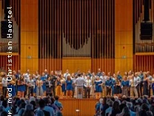 Ein Konzertchor steht auf einer Bühne in einem großen, gut besuchten Konzertsaal.