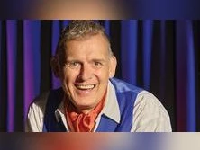 Ein Mann in Bühnenkostüm mit freundlichem Lächeln vor einem Vorhang in Blau- und Lila-Tönen.