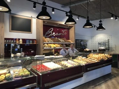brante.jpg Das Bild zeigt den Innenbereich der Bäckerei Brante mit einer Theke voller Backware, hinter der zwei Mitarbeiter stehen. Die Beleuchtung besteht aus mehreren schwarzen Hängelampen. Im Hintergrund ist ein Regal mit verschiedenen Broten sowie ein großes Schild mit den Namen "Brante" zu sehen.