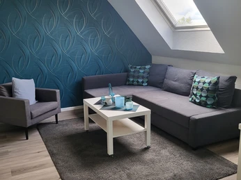 Wohnbereich in der Ferienwohnung Das Fischerhaus in Velbert Moderner Wohnbereich mit grauem Sofa und Sessel, blaue Akzentwand, großem Fenster, Teppich und Couchtisch.