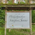 Holzschild mit Aufschrift "Heidschnucken-Schäferei Senne" vor grünem Wald im Hintergrund.