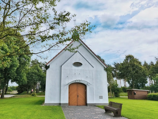 Weiße Kapelle mit Spitzdach vor Waldkulisse, hölzerne Doppeltür, gepflasterter Weg, Bank im Vordergrund.