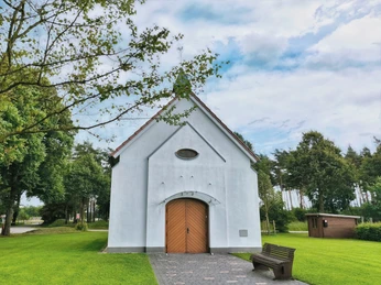 Hövelsenner Kapelle Weiße Kapelle mit Spitzdach vor Waldkulisse, hölzerne Doppeltür, gepflasterter Weg, Bank im Vordergrund.