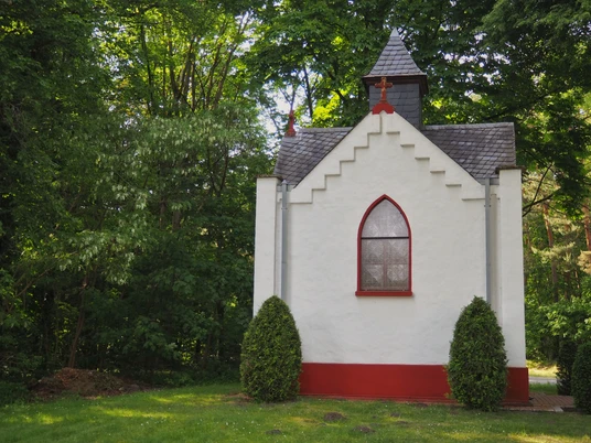 Kleine weiße Kapelle mit rotem Sockel, gotischem Fenster und grüner Umgebung in Hövelhof.