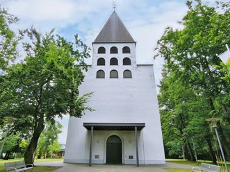 Weiße Kirche mit grauem Turm, sechs Bogenfenstern und grünem Umfeld durch Bäume flankiert.