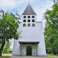 Herz-Jesu Kirche Hövelriege Weiße Kirche mit grauem Turm, sechs Bogenfenstern und grünem Umfeld durch Bäume flankiert.