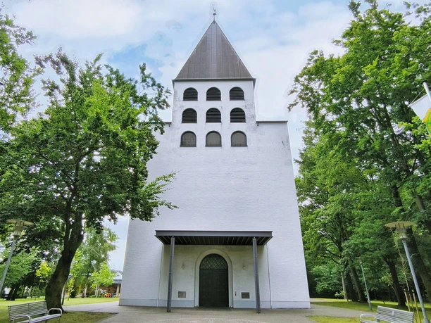Herz-Jesu Kirche Hövelriege Weiße Kirche mit grauem Turm, sechs Bogenfenstern und grünem Umfeld durch Bäume flankiert.