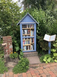 Bild Bücherschrank.jpeg Ein blauer Bücherschrank thront idyllisch zwischen üppigem Grün, mit bunt gemischten Büchern und gepflastertem Boden davor.