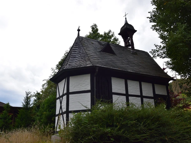 Barbarakapelle Fachwerk-Kapelle in Rösrath, mit Dachreiter und Glocke, umgeben von Bäumen und hohem Gras.
