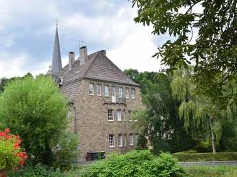 Ensemble Volberg Altes Steinhaus mit mehreren Schornsteinen und einem Kirchturm im Hintergrund, umgeben von grüner Vegetation.