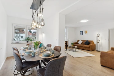 WohnLiebe_Luisenstrasse1.jpeg Modernes Wohnzimmer mit Esstisch und acht Stühlen. Lampen hängen von der Decke, zwei Sofas im Hintergrund.