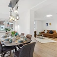 WohnLiebe_Luisenstrasse1.jpeg Modernes Wohnzimmer mit Esstisch und acht Stühlen. Lampen hängen von der Decke, zwei Sofas im Hintergrund.