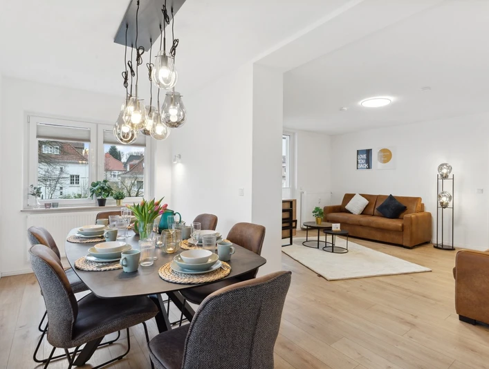 WohnLiebe_Luisenstrasse1.jpeg Modernes Wohnzimmer mit Esstisch und acht Stühlen. Lampen hängen von der Decke, zwei Sofas im Hintergrund.