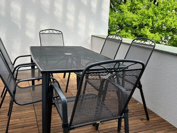 WohnLiebe_Luisenstrasse10.jpeg Sechs Metallstühle um einen großen Gartentisch auf einer Balkonfläche mit Holzboden und weißer Mauer.