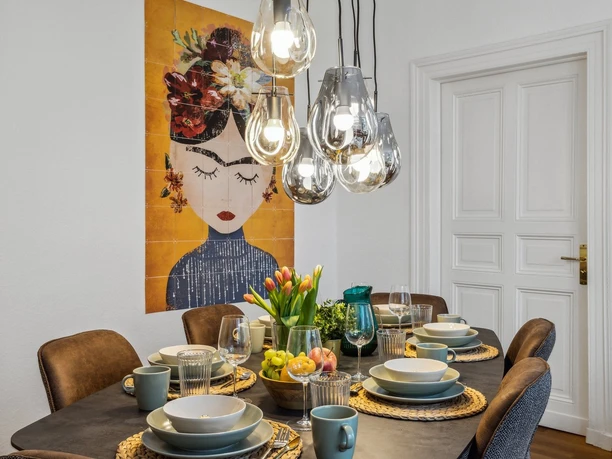 WohnLiebe_Augustaplatz1.jpeg Ein stilvoll gedeckter Esstisch mit moderner Beleuchtung, buntem Blumenarrangement und Kunst an der Wand.