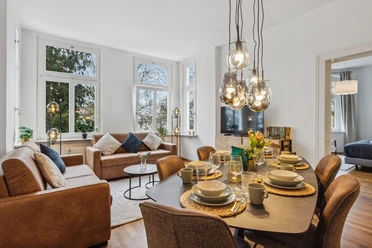 WohnLiebe_Augustaplatz2.jpeg Modernes Wohnzimmer mit großen Fenstern, bequemen Sofas, Esstisch und stilvollen Hängeleuchten.