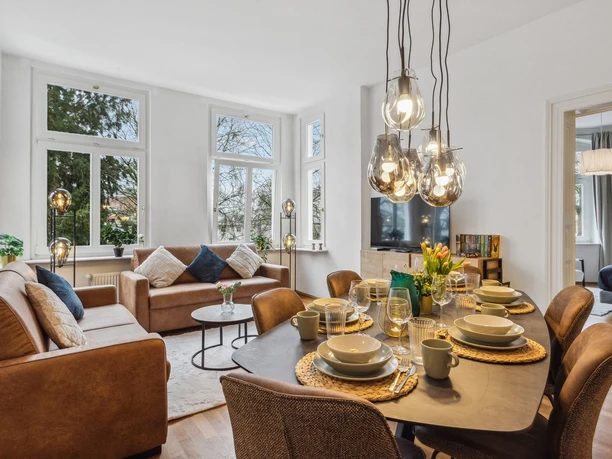 WohnLiebe_Augustaplatz2.jpeg Modernes Wohnzimmer mit großen Fenstern, bequemen Sofas, Esstisch und stilvollen Hängeleuchten.